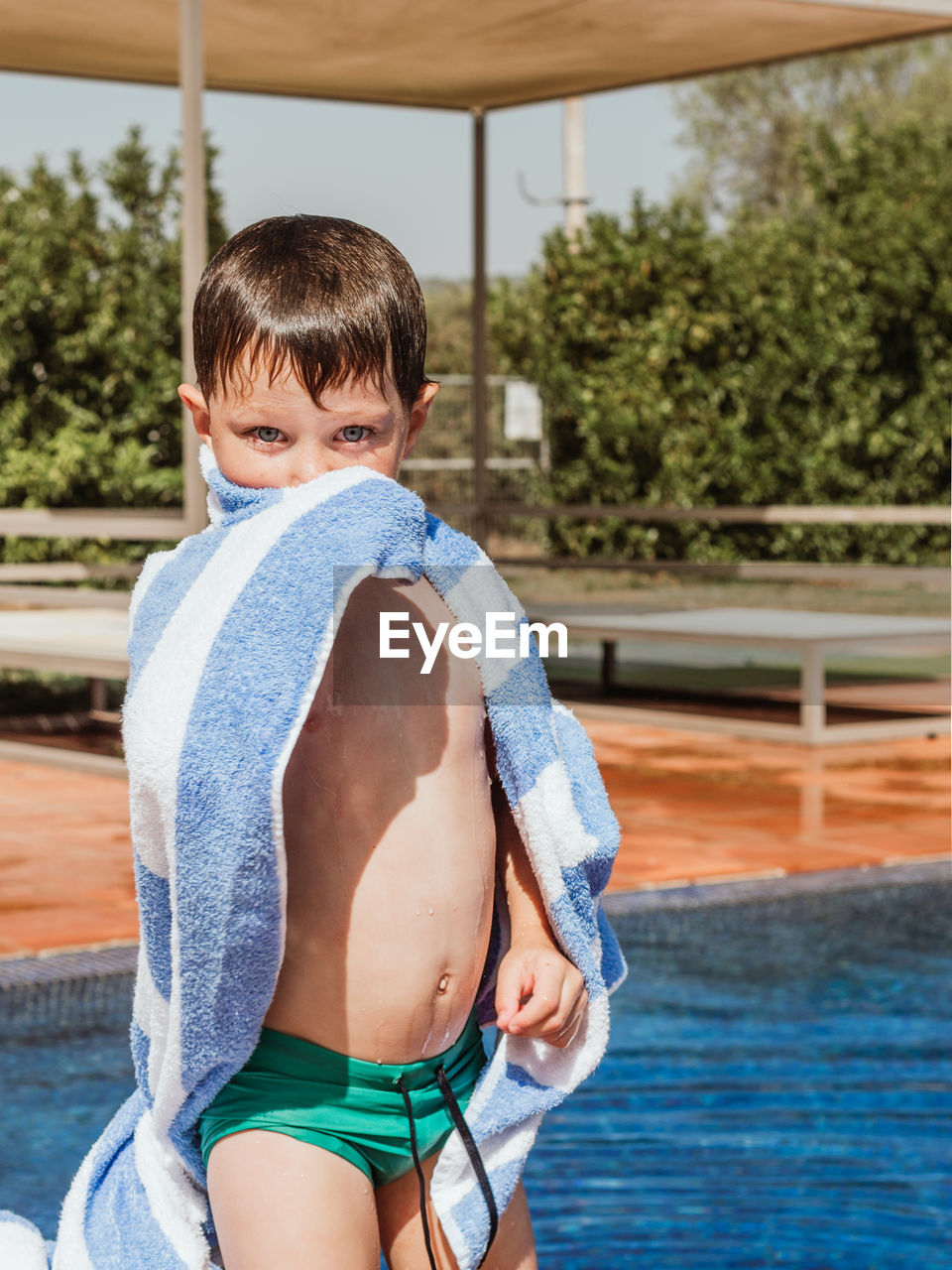 Adorable little kid with wet hair wiping body with towel while standing near swimming pool on sunny day in summer and looking at camera