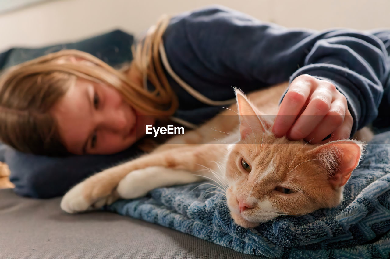 A young adolescent girl lying on a couch finds comfort by snuggling and petting her tabby cat