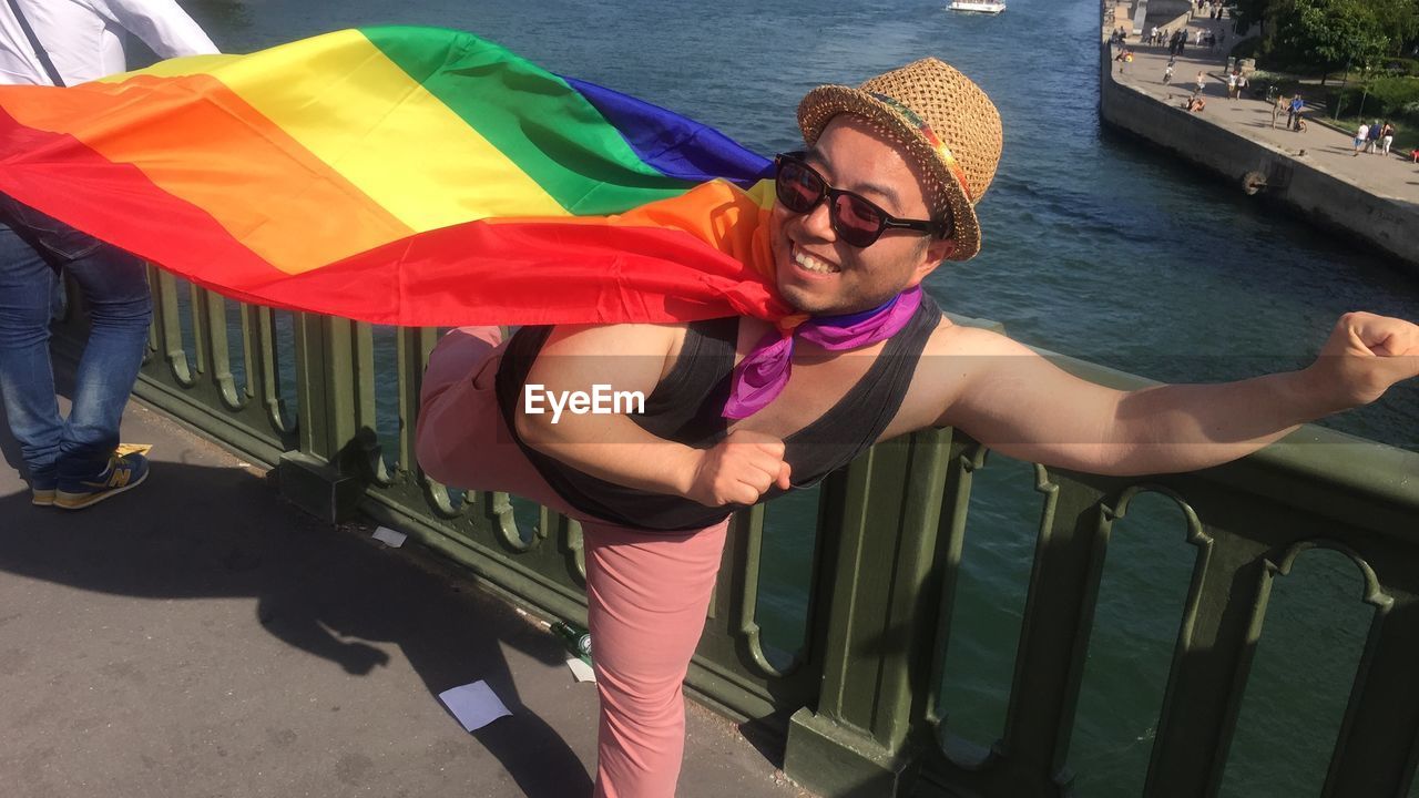 Cheerful man wearing rainbow flag by railing against sea