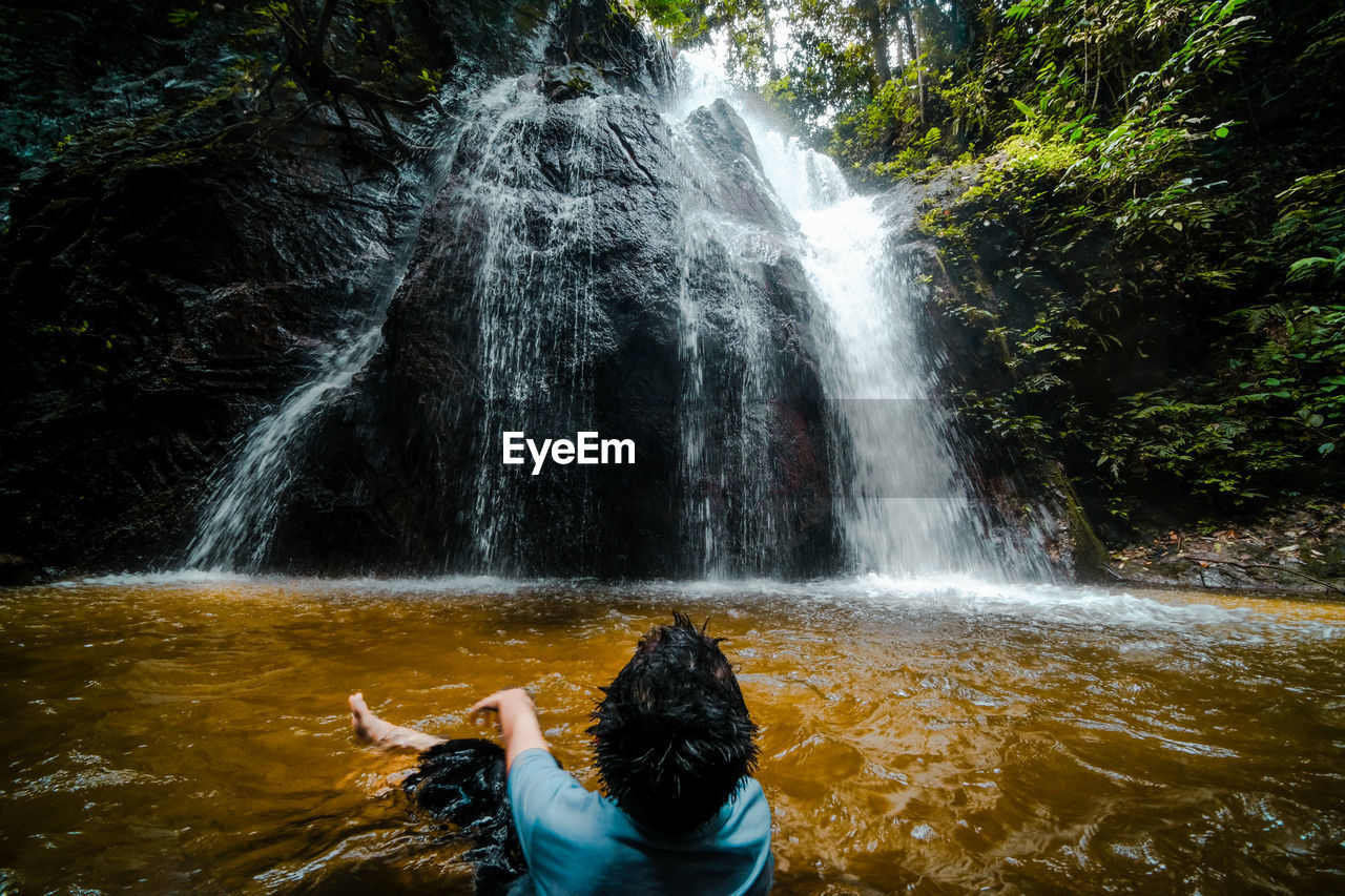 Rear view of boy sitting in water against waterfall 