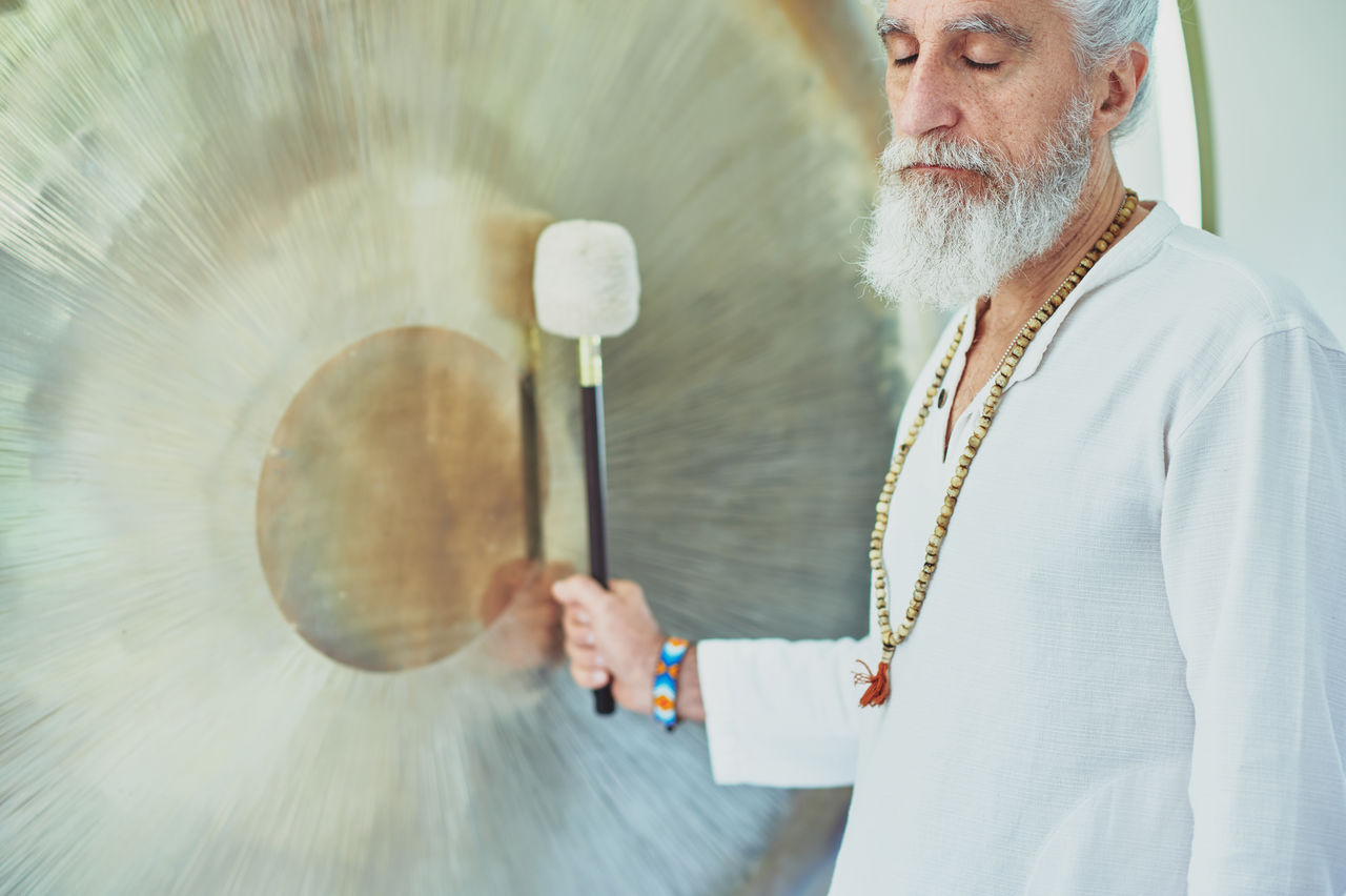 Side view of crop adult male in white clothes with mallet playing suspended gong during spiritual practice