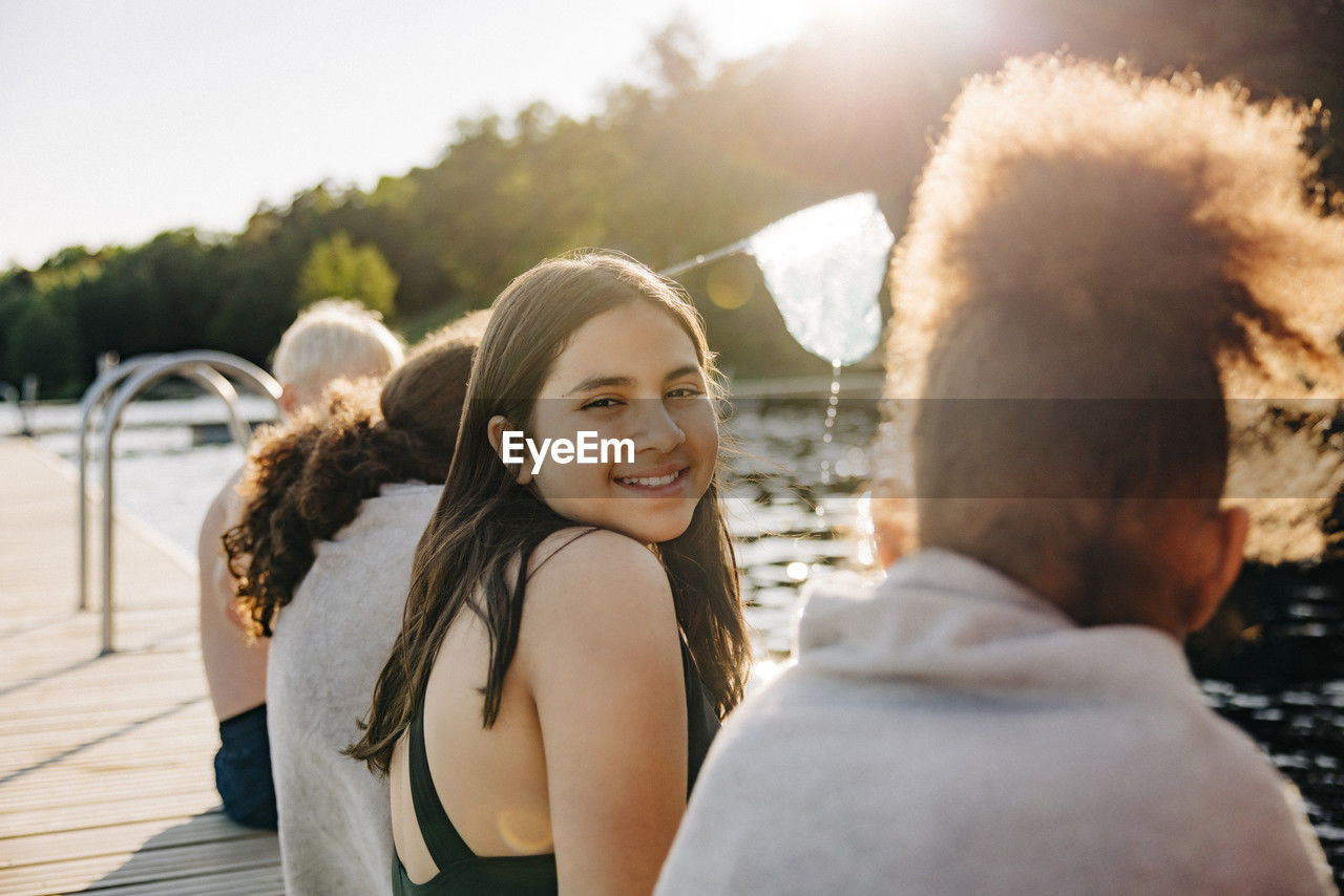 Portrait of smiling girl sitting with friends on jetty by lake