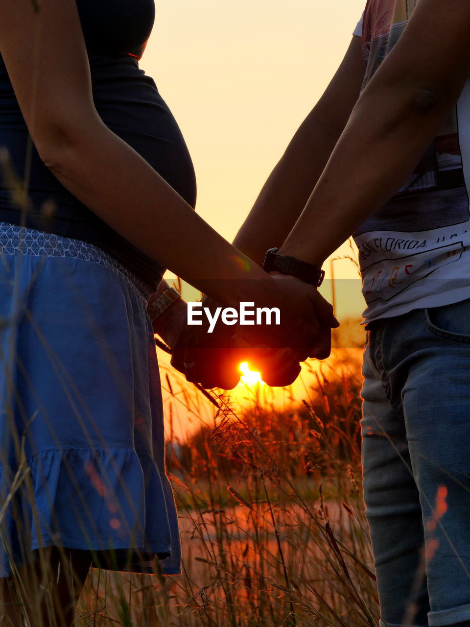 Midsection of couple holding hands against sky at sunset