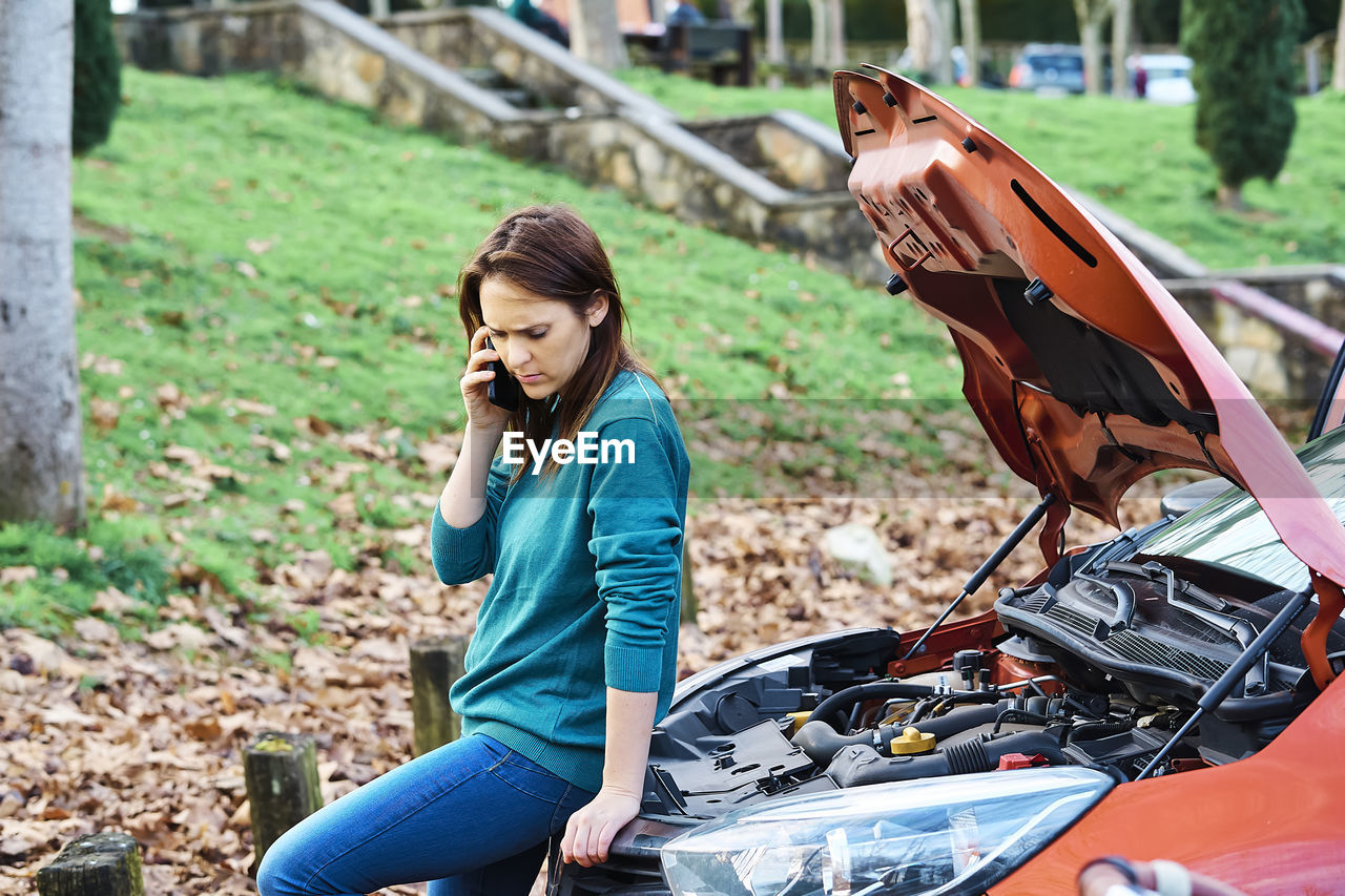 YOUNG WOMAN SITTING ON CAR BY OUTDOORS