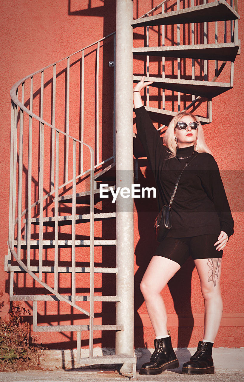 FULL LENGTH PORTRAIT OF YOUNG WOMAN STANDING BY RAILING AGAINST BUILDING