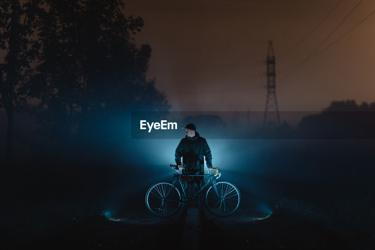 Man with bicycle against illuminated light standing on road at night