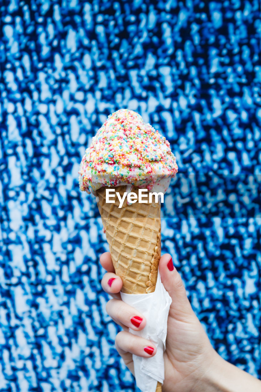 Cropped hand of woman holding sprinkles ice cream cone against blue wall