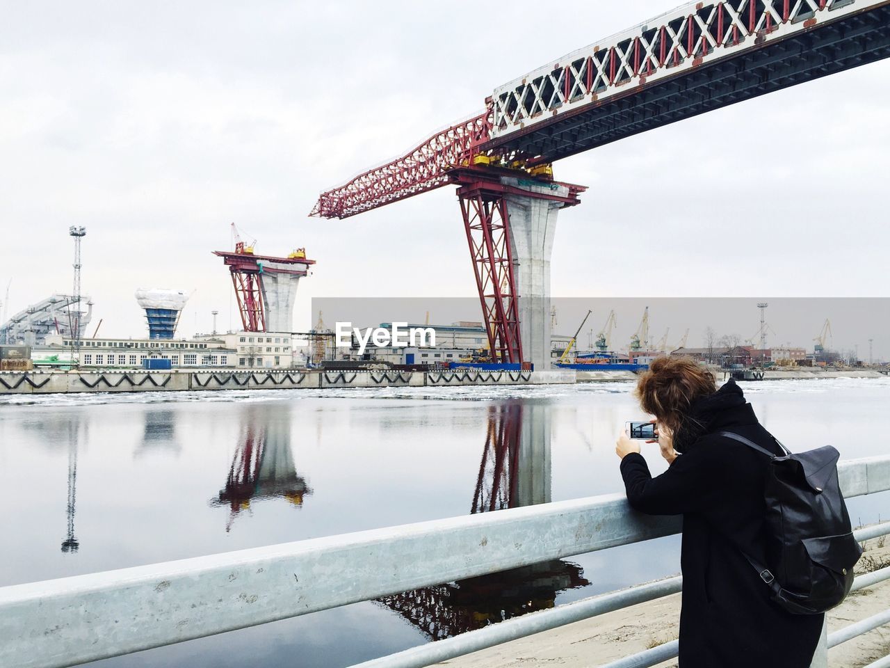 Woman photographing incomplete bridge over river in city against sky