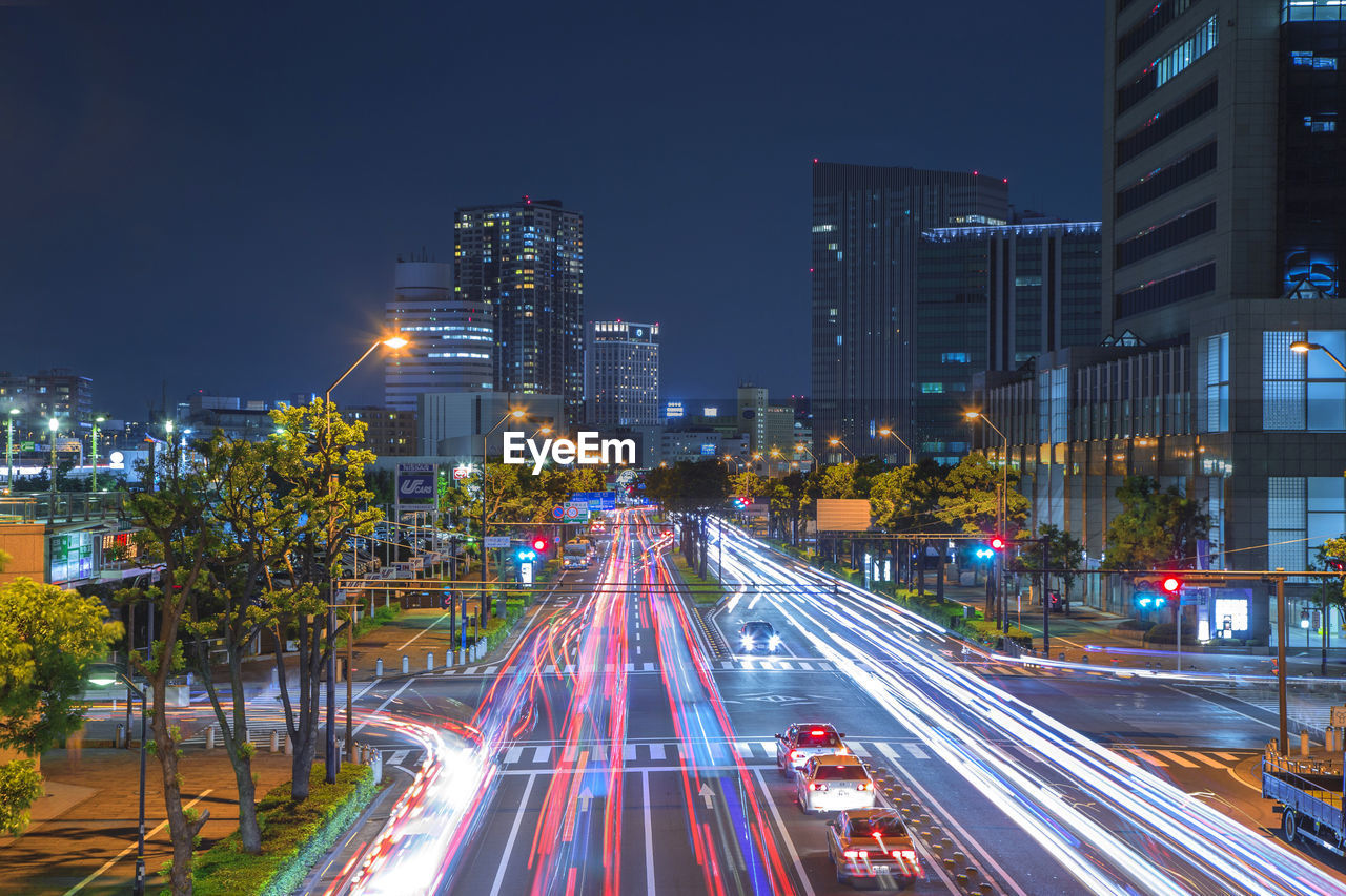 Light trails on city street by buildings against sky at night