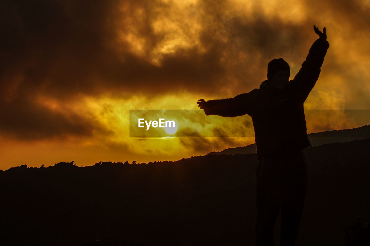 Silhouette man with arms outstretched standing against orange sky