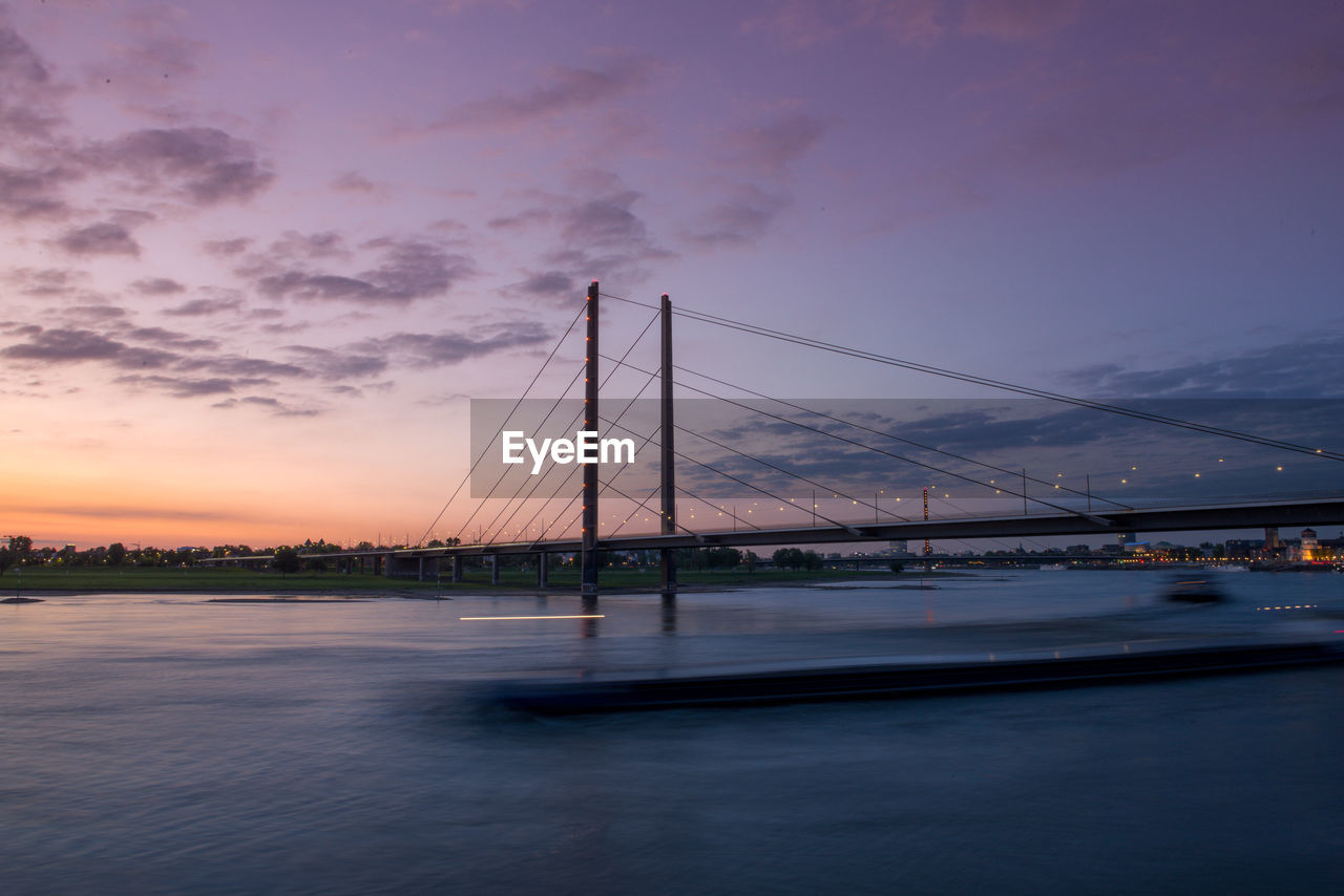 Long exposure of a bridge over river against sky during sunset with passing barge