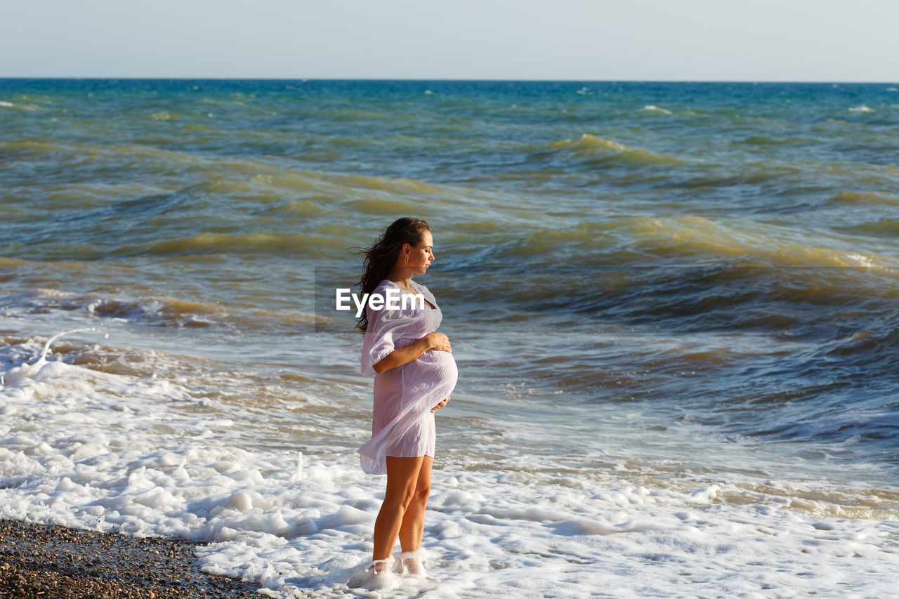 Portrait beautiful young pregnant woman, against blue sea on beach, thoughtful