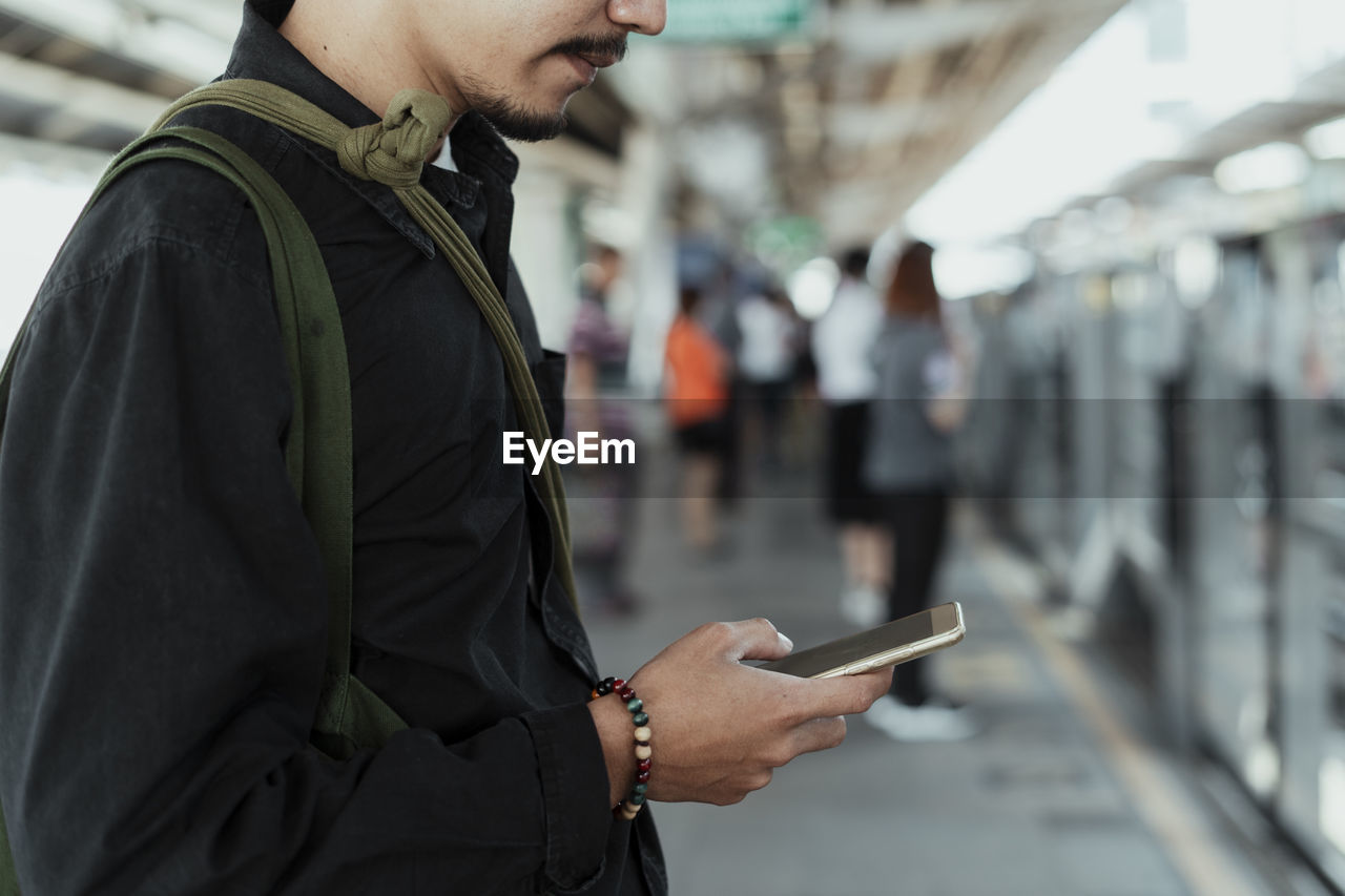 Midsection of man using smart phone while standing at railroad platform