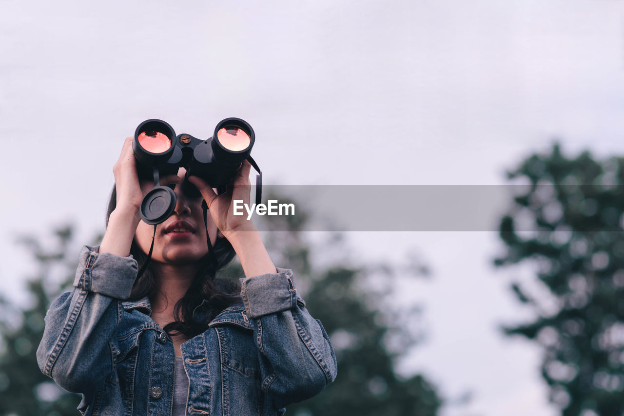 Young woman looking through binoculars while standing against sky