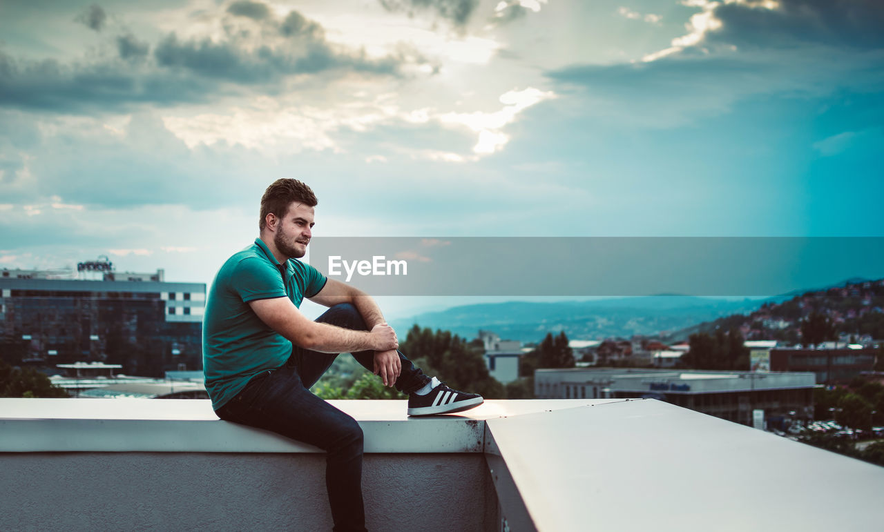 FULL LENGTH OF YOUNG MAN SITTING IN CITY AGAINST SKY