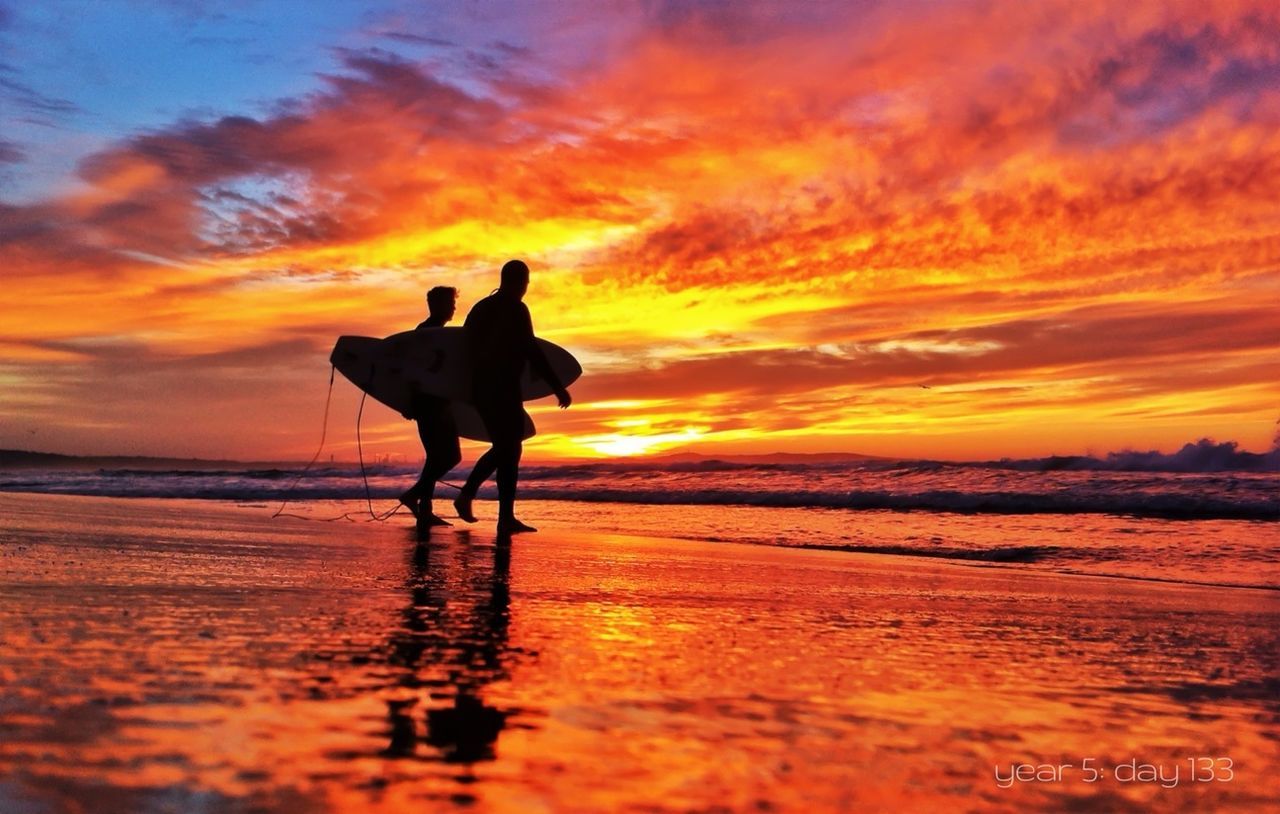 Two silhouette people with surfboards walking on beach at sunset