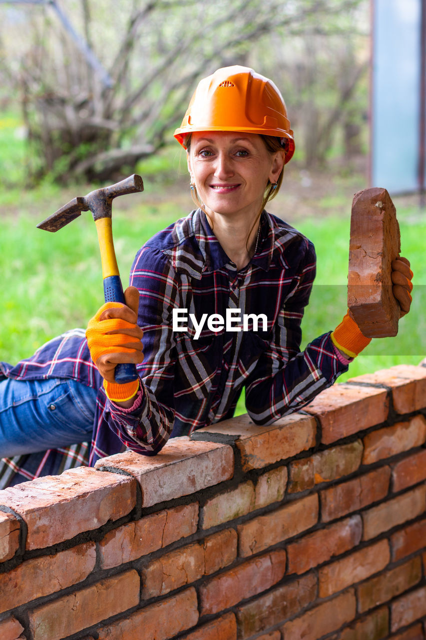 A young woman in a construction helmet with a hammer and a brick in her hands smiles