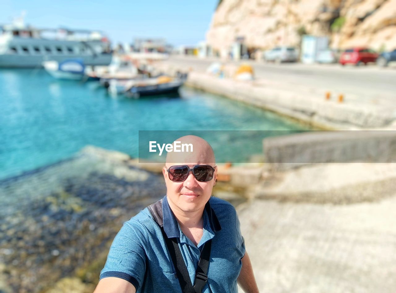 sunglasses, one person, portrait, glasses, water, adult, fashion, vacation, smiling, looking at camera, day, architecture, front view, nature, happiness, men, travel destinations, lifestyles, sea, leisure activity, emotion, focus on foreground, casual clothing, travel, transportation, sunlight, outdoors, holiday, trip, city, relaxation, mature adult, headshot, tourism, summer, sunny, person, clothing, tourist, waist up, standing, nautical vessel, goggles, enjoyment, cool attitude, sky, copy space, cheerful