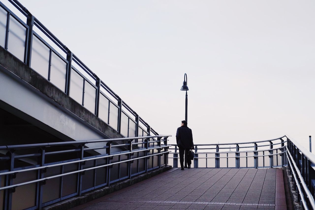 MAN STANDING ON FOOTBRIDGE AGAINST CLEAR SKY