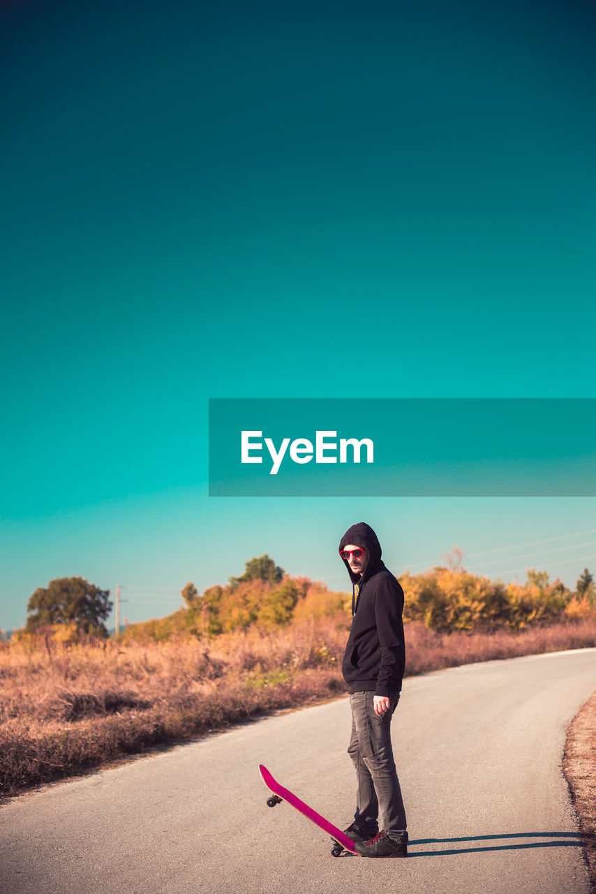 Side view of man with skateboard standing on road against clear blue sky