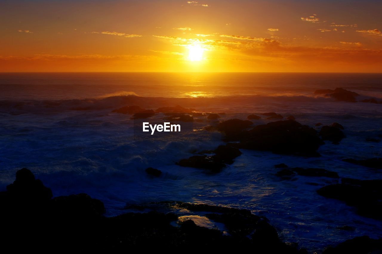 Scenic view of sunset with silhouette rocks in sea against orange sky