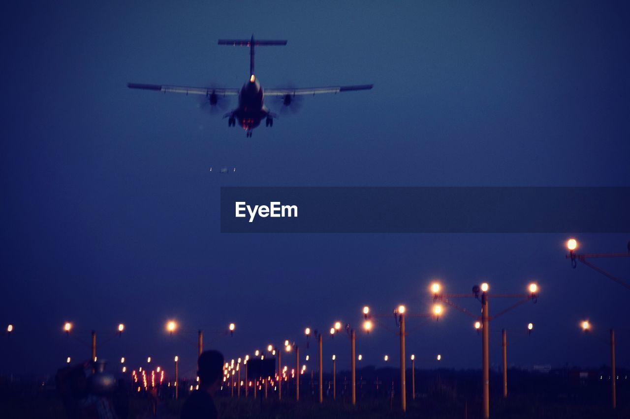 Low angle view of airplane flying over illuminated runway against clear blue sky