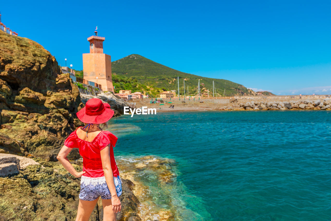 Rear view of woman standing on rock formations by sea against blue sky