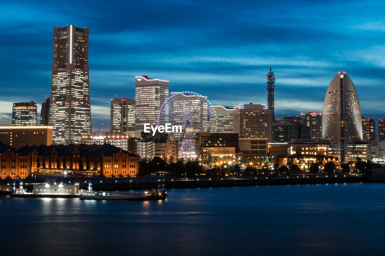 Illuminated modern buildings by river against sky in city