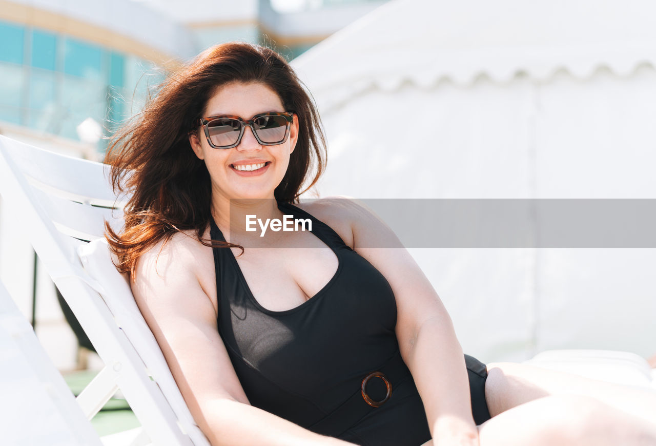 Stylish woman plus size body positive in black swimsuit and sunglasses on beach lounger near pool