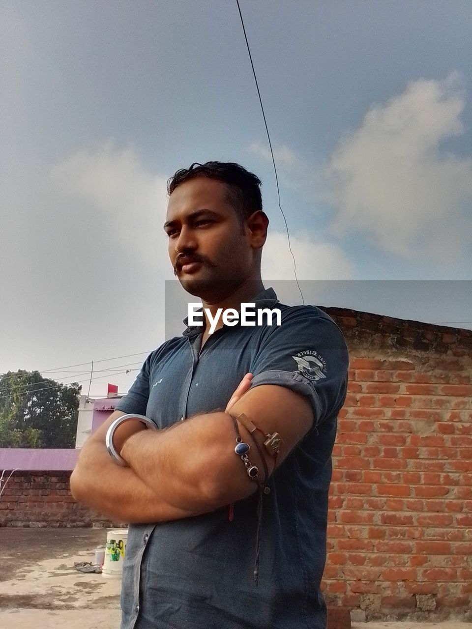 one person, standing, sky, adult, men, young adult, arms crossed, architecture, cloud, waist up, lifestyles, casual clothing, serious, portrait, looking, looking away, day, person, leisure activity, nature, sports, outdoors, city, clothing, brick, blue, built structure, building exterior, low angle view
