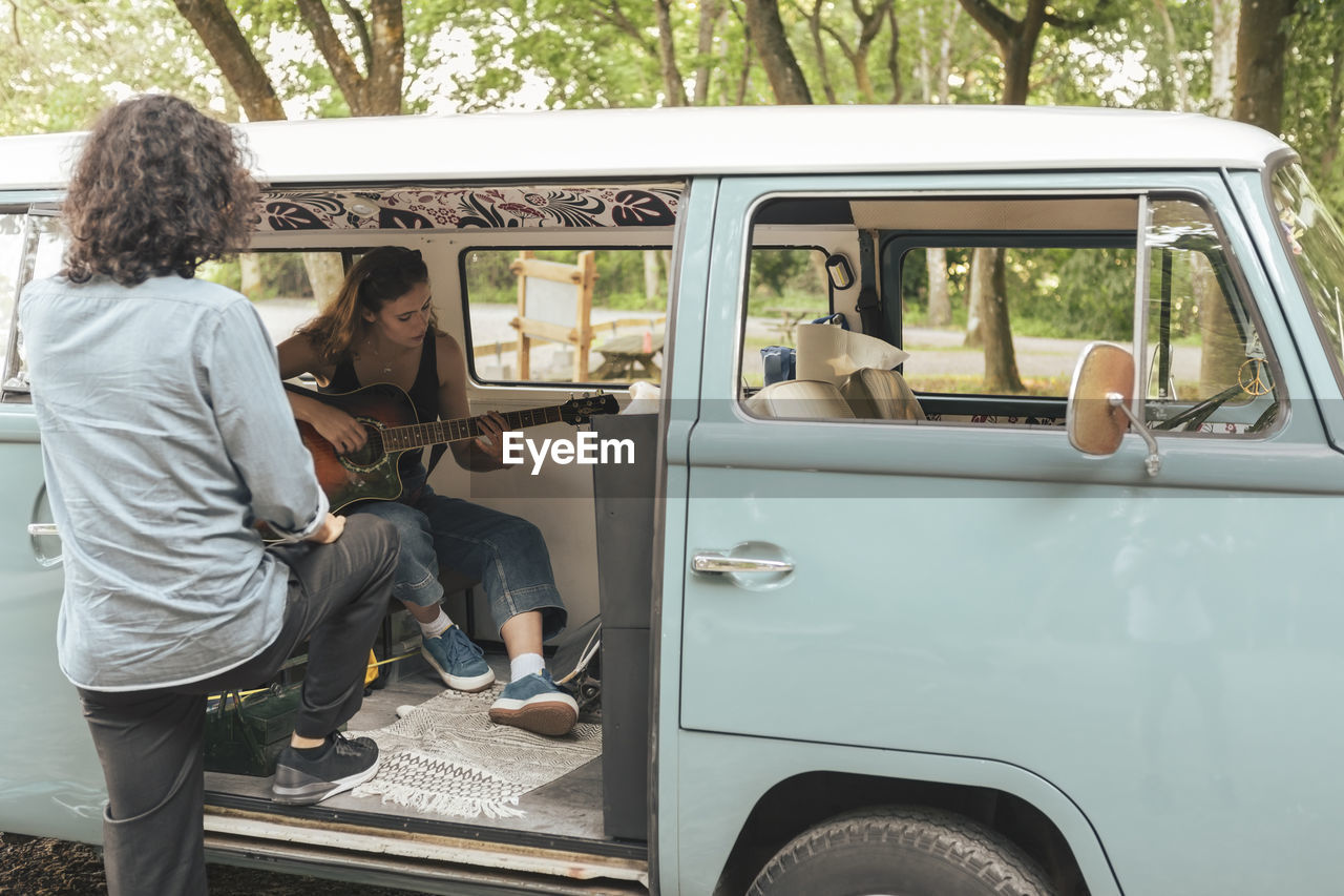 Rear view of man looking at female friend playing guitar inside van