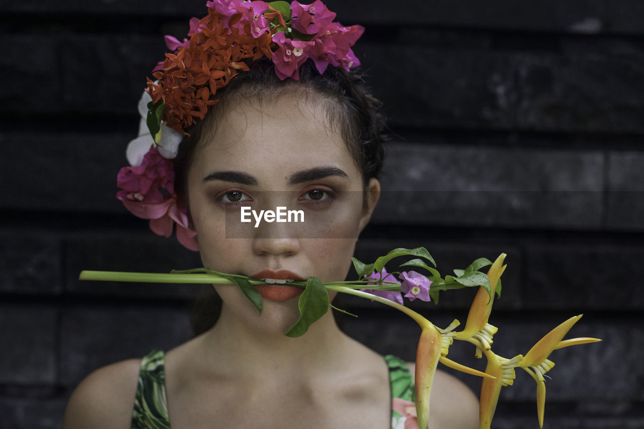 Portrait of young woman with flower in mouth standing against wall