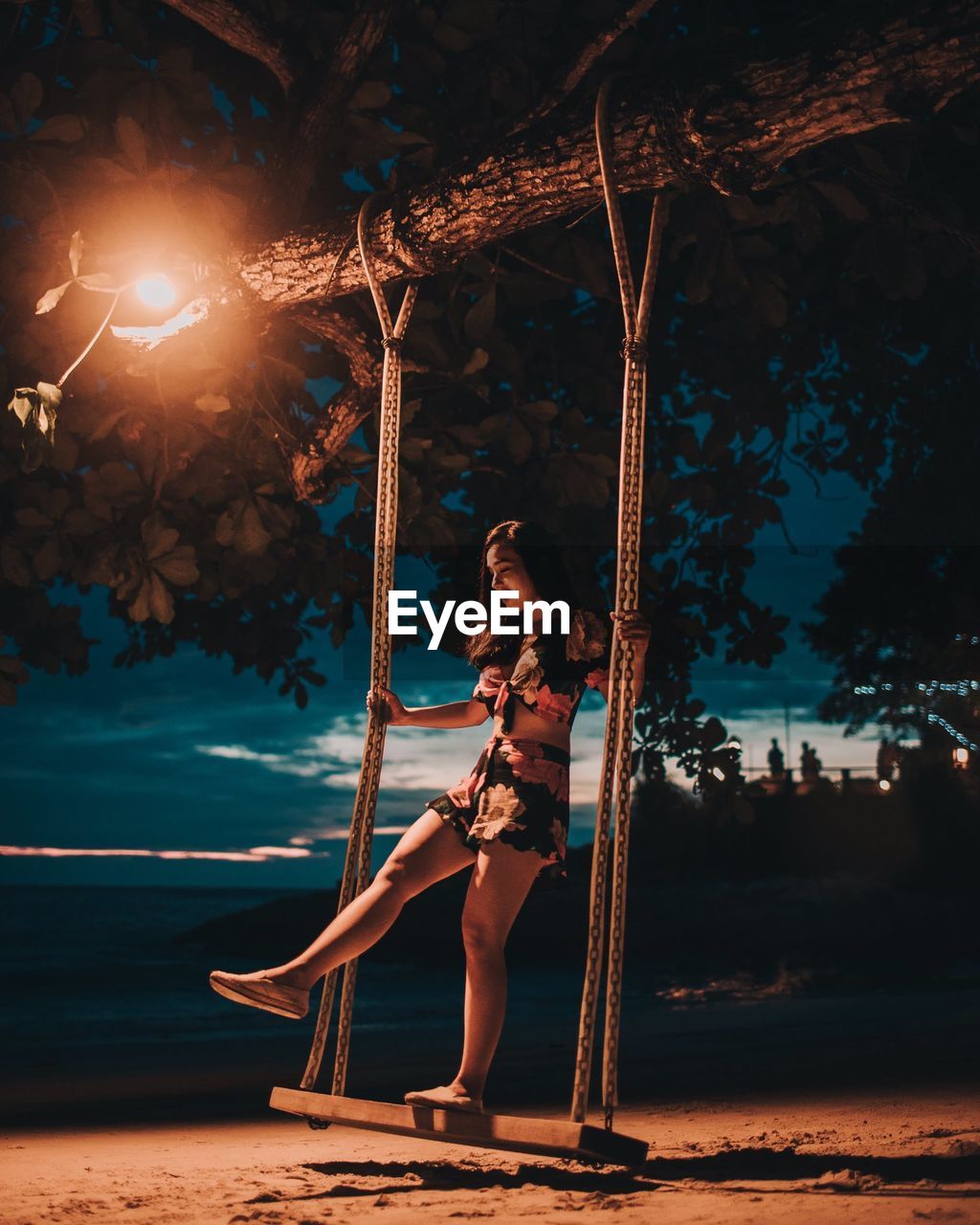 BEAUTIFUL YOUNG WOMAN SITTING ON TREE AT BEACH AGAINST SKY