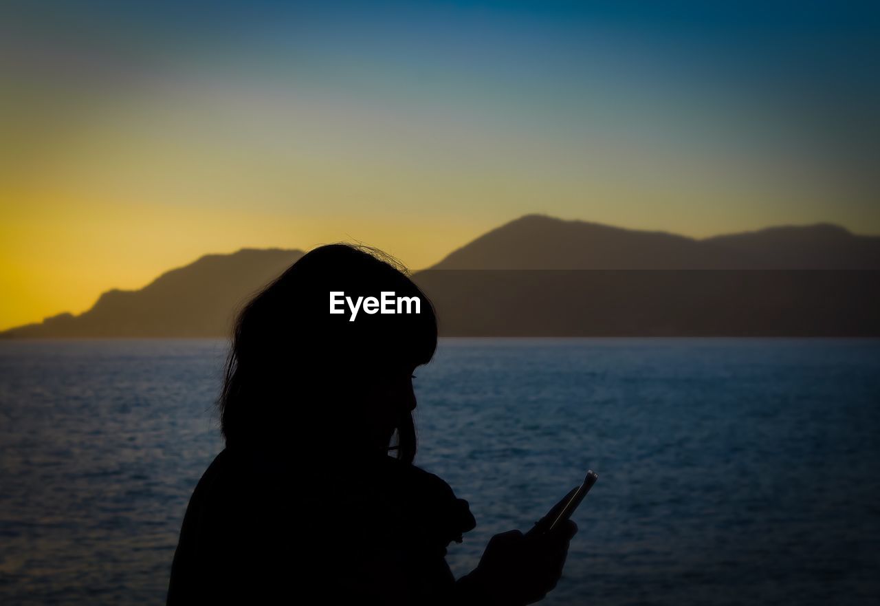 Silhouette woman using mobile phone against sea during sunset