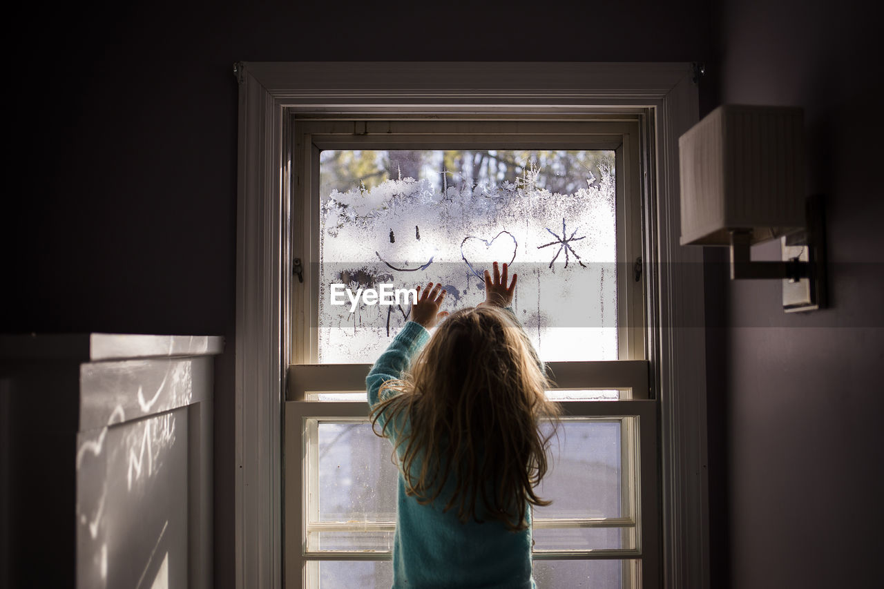 Rear view of girl drawing on condensed window at home