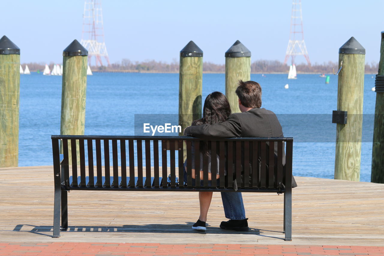 Rear view of couple sitting at riverbank against clear sky