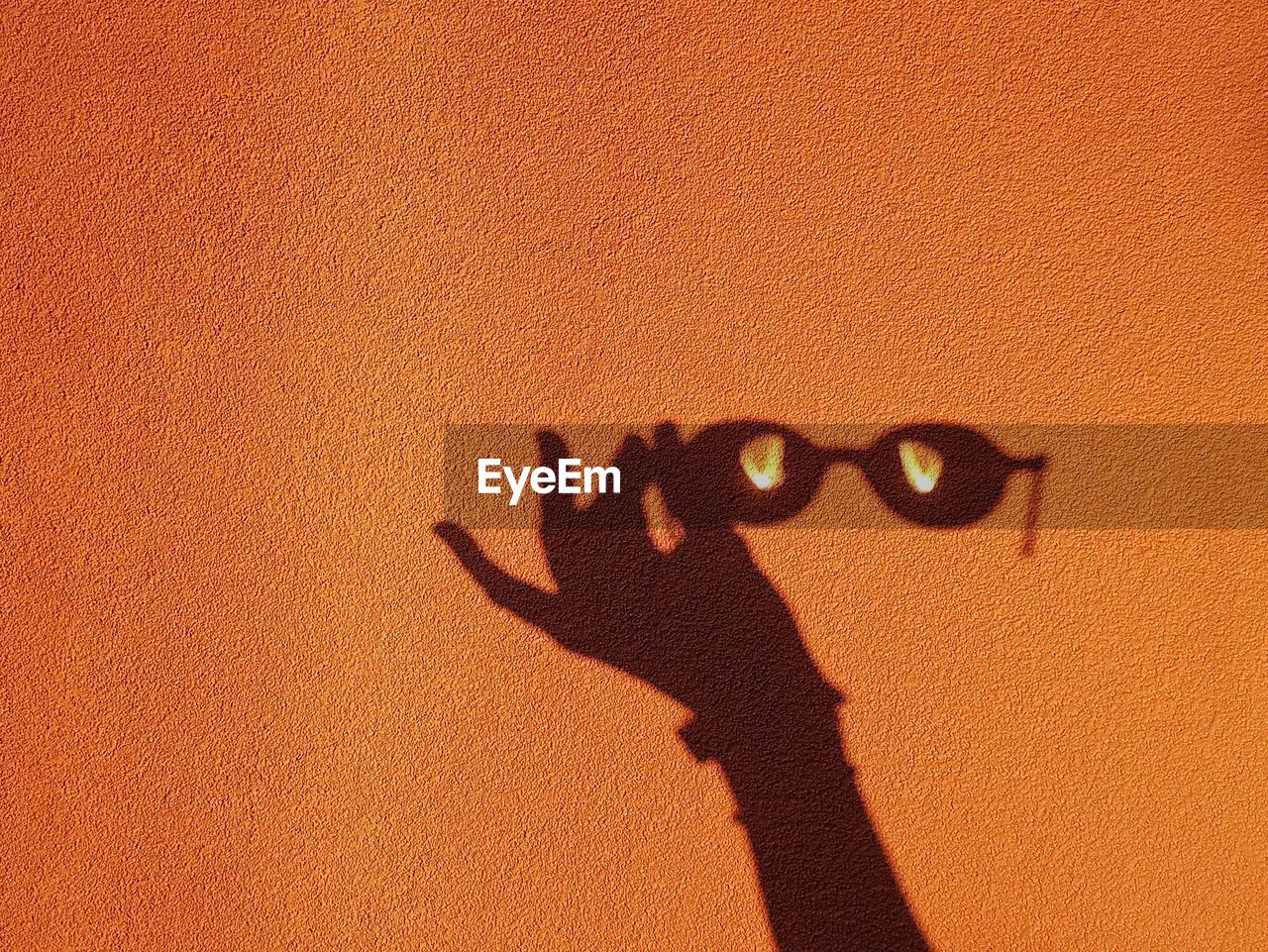 Shadow of hand holding sunglasses on orange wall during sunny day