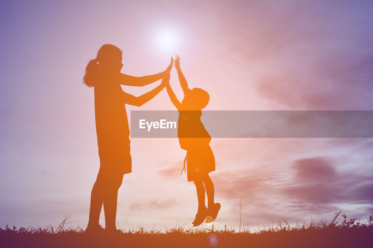 Silhouette siblings giving high-five against sky during sunset