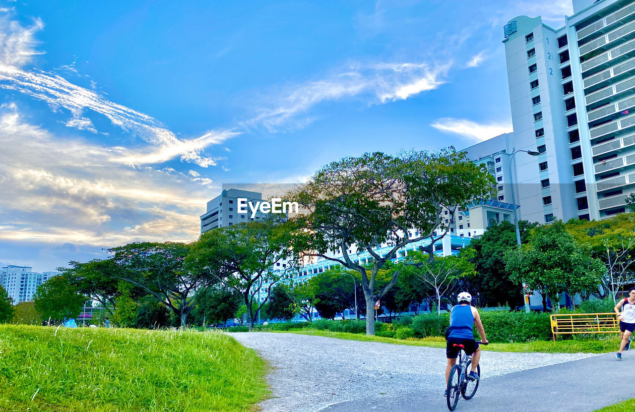 REAR VIEW OF MAN RIDING BICYCLE BY BUILDING IN CITY AGAINST SKY