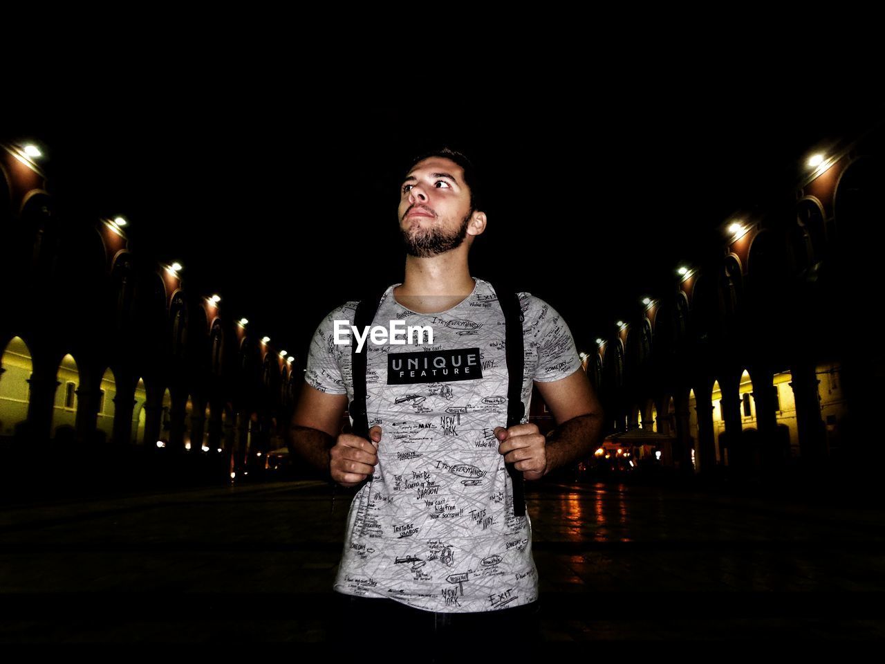 YOUNG MAN LOOKING AWAY WHILE STANDING AGAINST ILLUMINATED LIGHTS