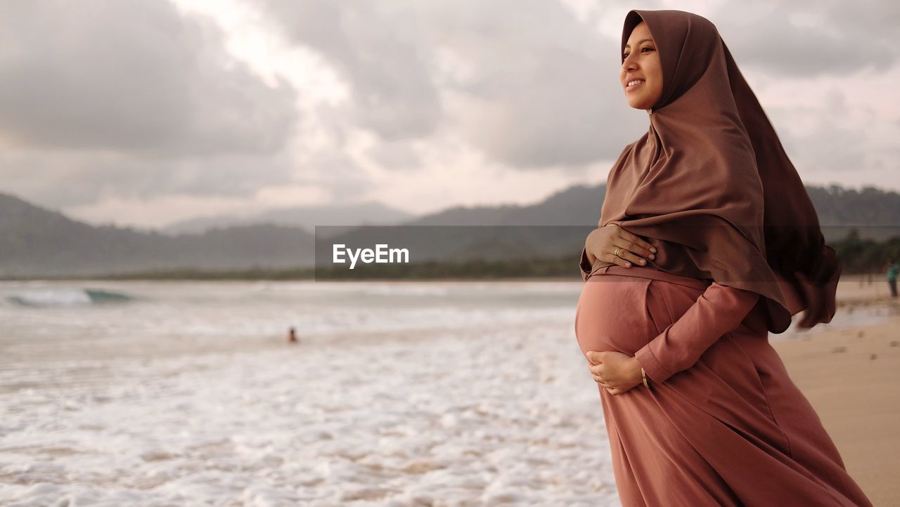 Smiling pregnant woman with hands on stomach wearing hijab standing at beach against cloudy sky