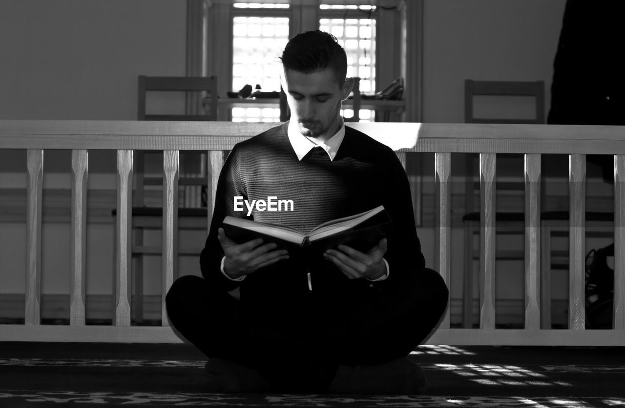 Full length of concentrated young man reading book while sitting against railing