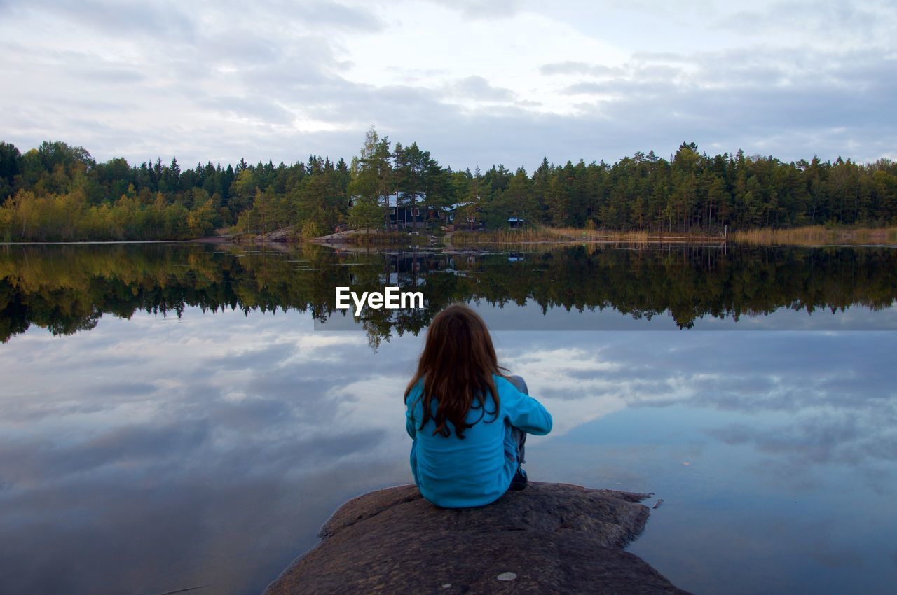 Rear view of girl looking at lake while sitting on rock in forest