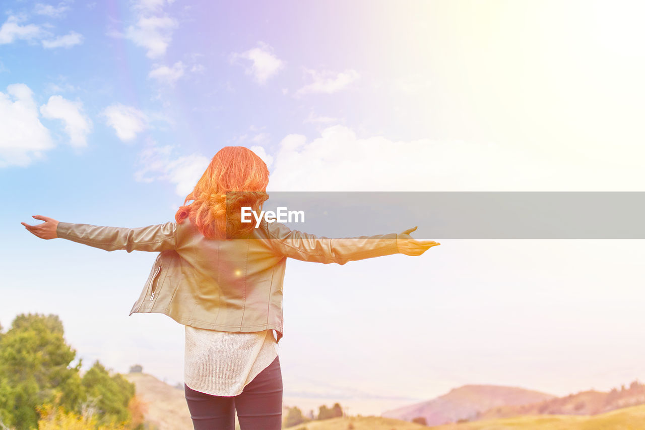 REAR VIEW OF WOMAN WITH ARMS OUTSTRETCHED AGAINST SKY