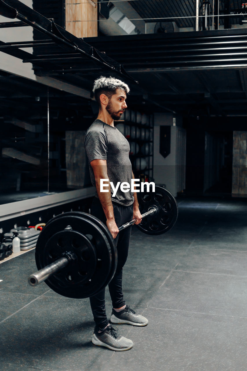 Full body side view of concentrated young bearded male athlete lifting heavy barbell during intense workout in contemporary sports club
