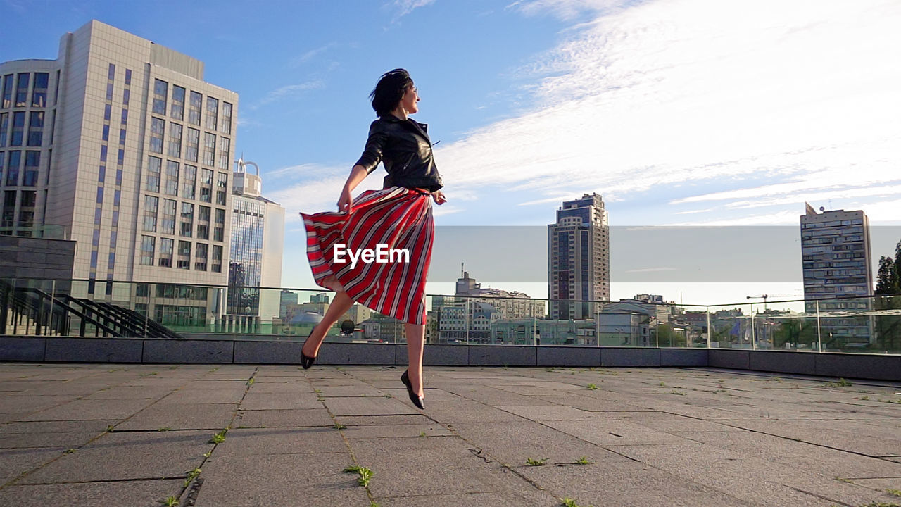 Happy woman wearing striped skirt and jacket on footpath in city