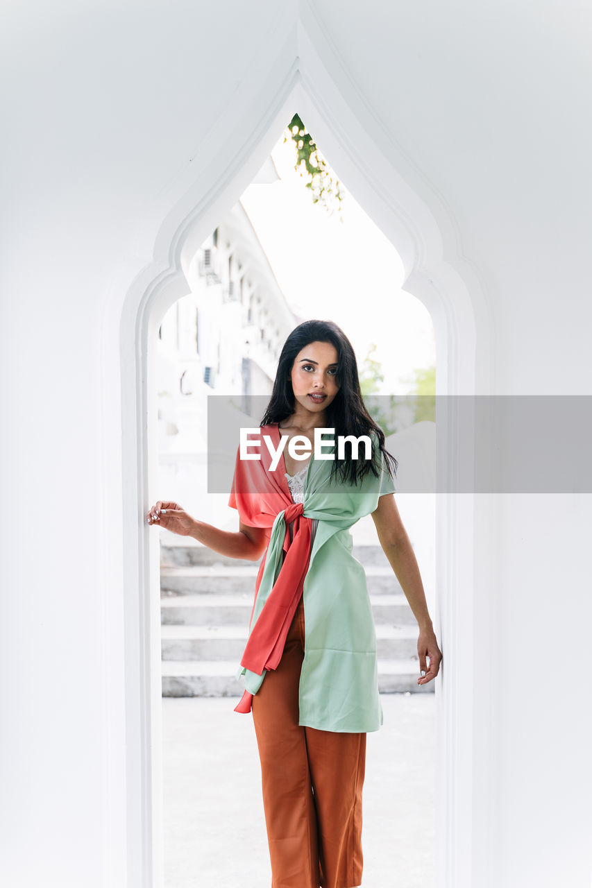 Cheerful charming young ethnic indian female in stylish clothes standing near on windowsill of arched window in oriental style house and looking at camera