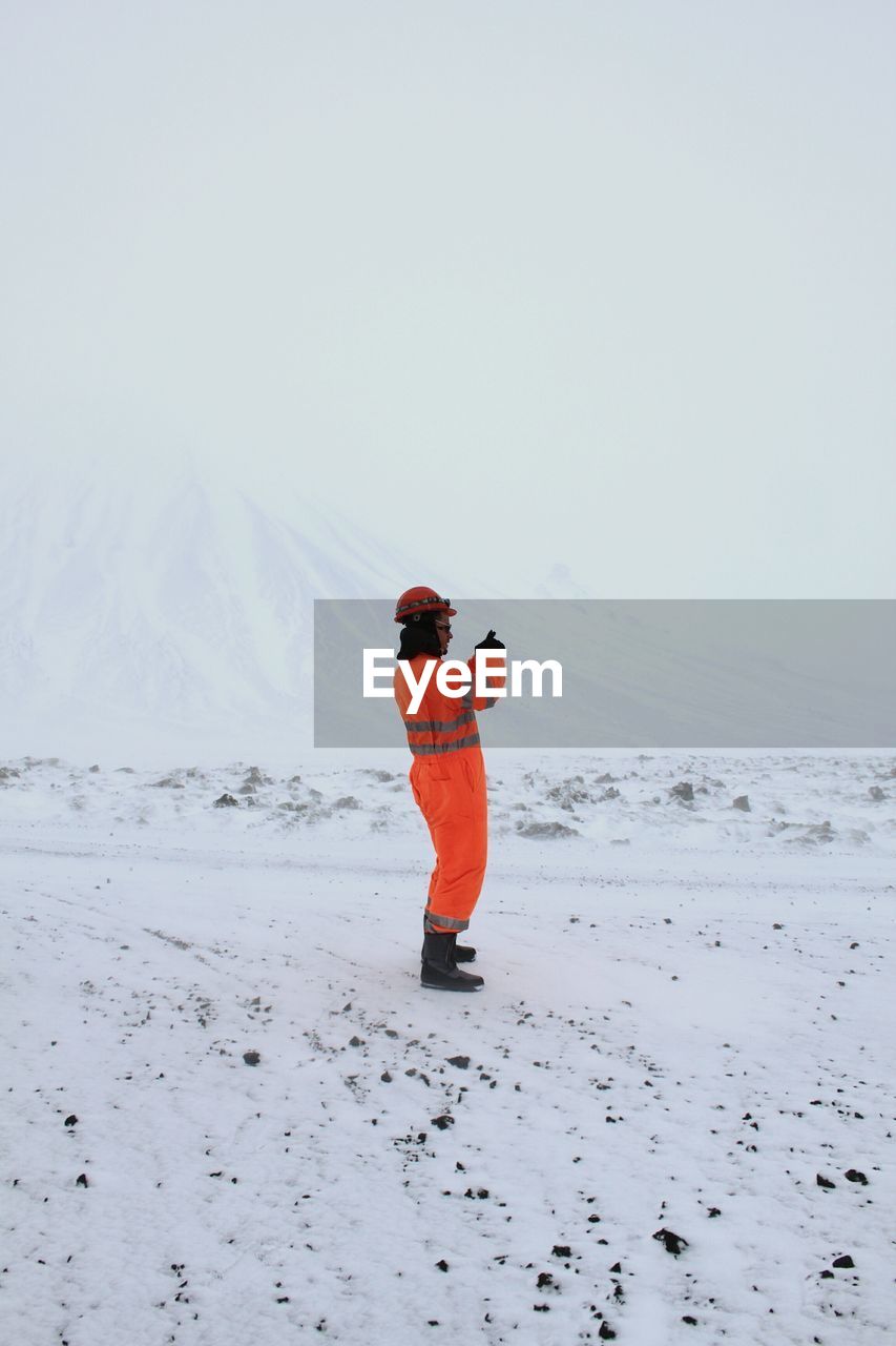 Side view of man wearing protective workwear standing on snow against clear sky