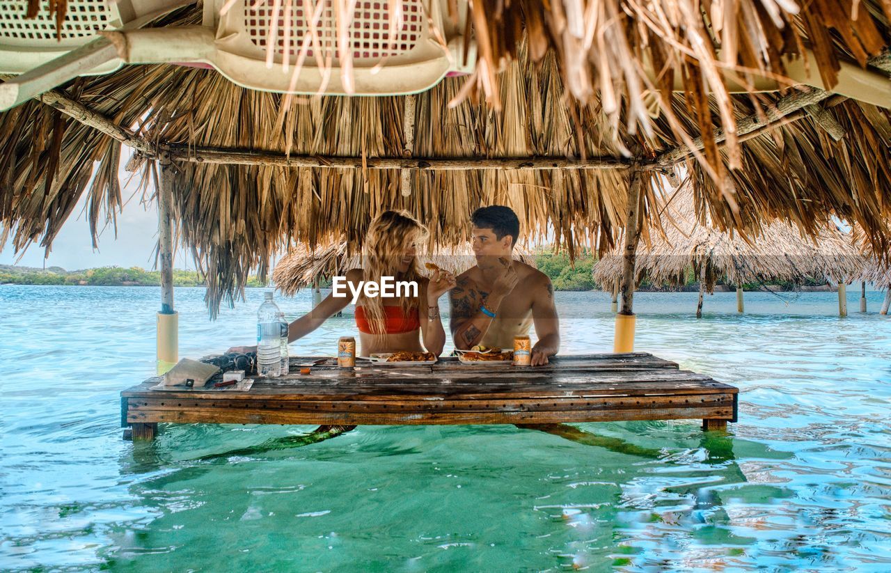 Friends having meal under thatched roof amidst sea