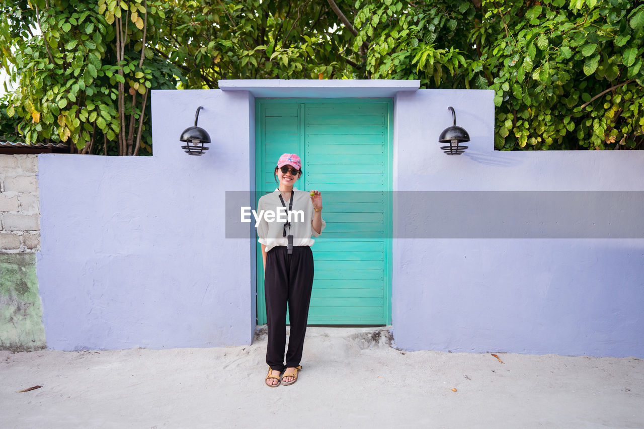 Full body happy young asian female tourist standing near colorful door and waving hand while spending summer holidays on maldives islands